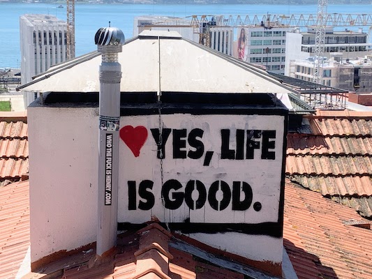 A sign painted above the roof of a building below the Miradouro de Santa Catarina. It says "Yes, Life is Good".