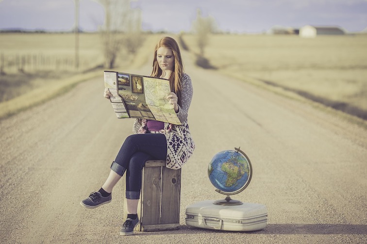 A woman sitting on a suitcase in the middle of a road, reading a map, with a globe next to her.