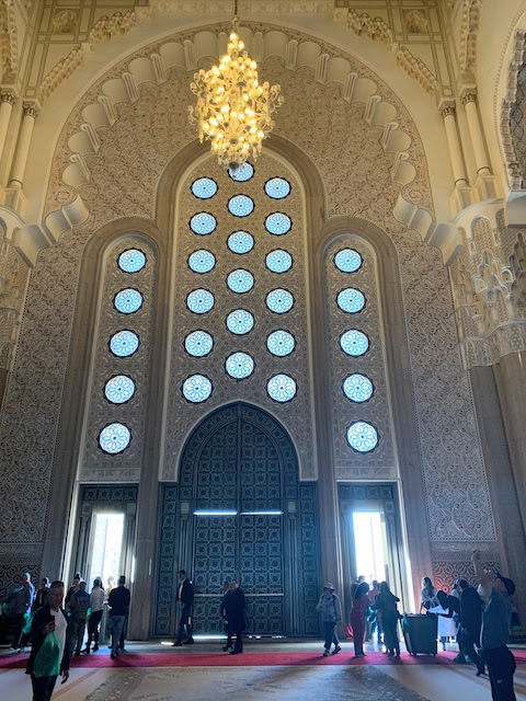 The interior of the Hassan II mosque showing large doors and a tall wall of decorative windows behind a chandelier.