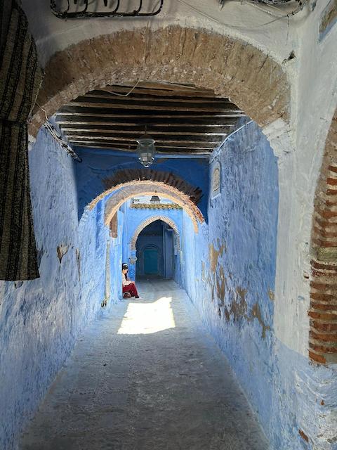 A long corridor in Chefchaouen with walls painted the traditional blue color that contrasts with brown brickwork and wooden beams in the ceiling.