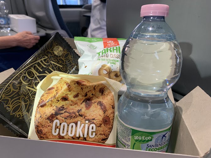 A snack box with a cookie, a bag of pretzels, a bottle of water and a moist towlette.