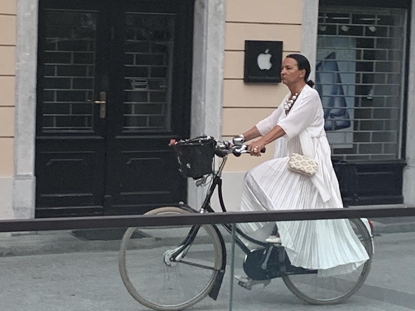 A woman wearing a flowing white dress arriving at the concert venue on a bicycle.