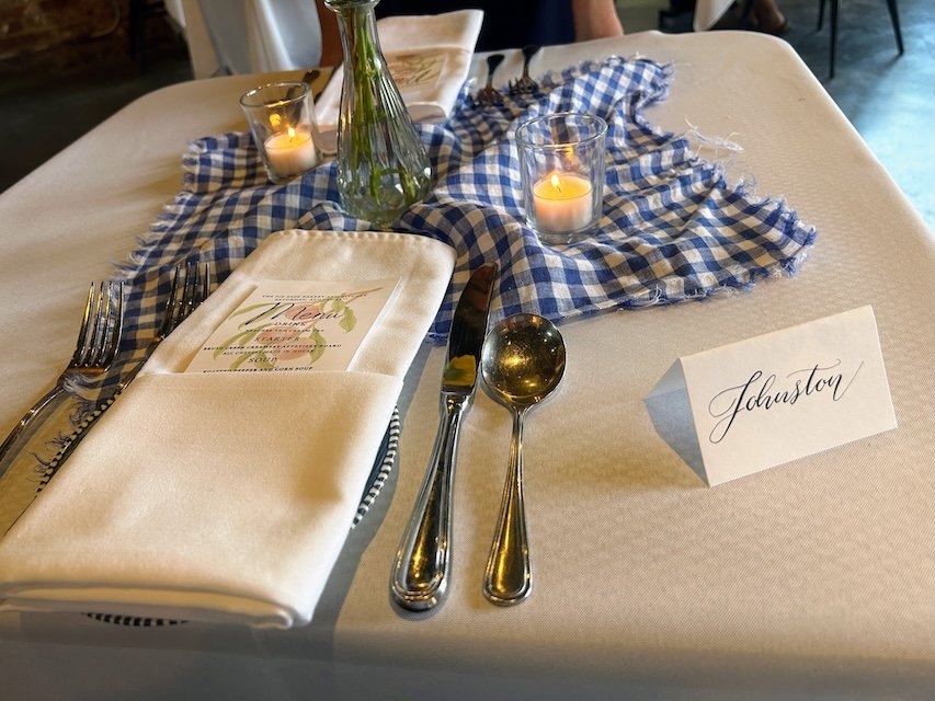 The table setting at the Special Dinner - a gingham cloth arranged casually on the table, two small candles, a place card with "Johnston" written in elaborate script, the menu tucked in the folds of the linen napkin, a small vase of flowers in the center.
