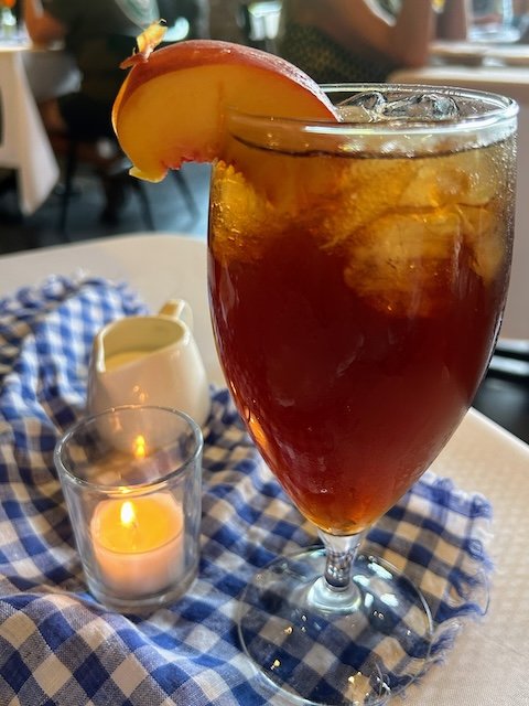 A glass of peach iced tea, served with a wedge of peach on the glass and a small pitcher of cream to add to the tea.